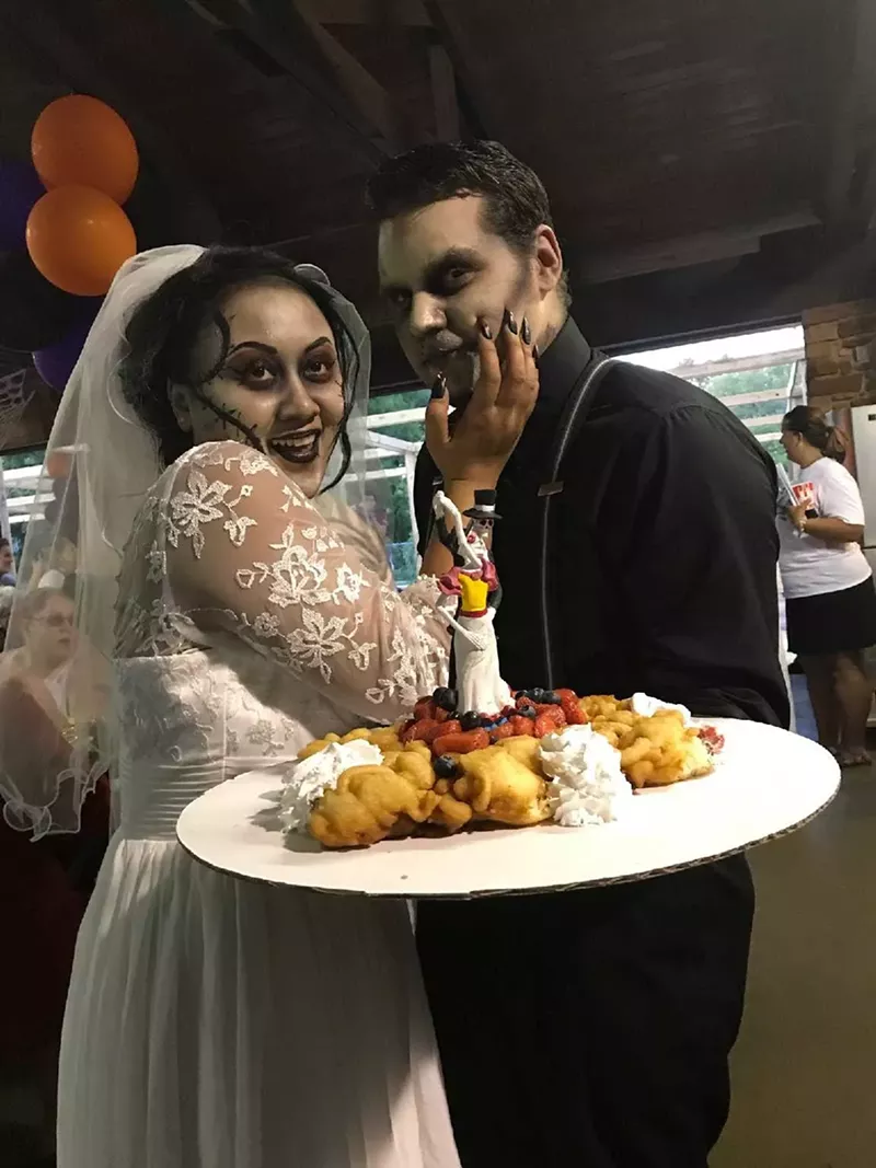 Image: Ethan and Chantra Briggs prepare to share a bite of their funnel wedding cake at a Halloween-themed wedding ceremony at Six Flags Over Texas.