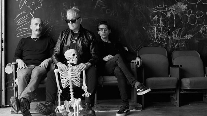 The band members of Alkaline Trio pose with a skeleton in a black and white press photo.