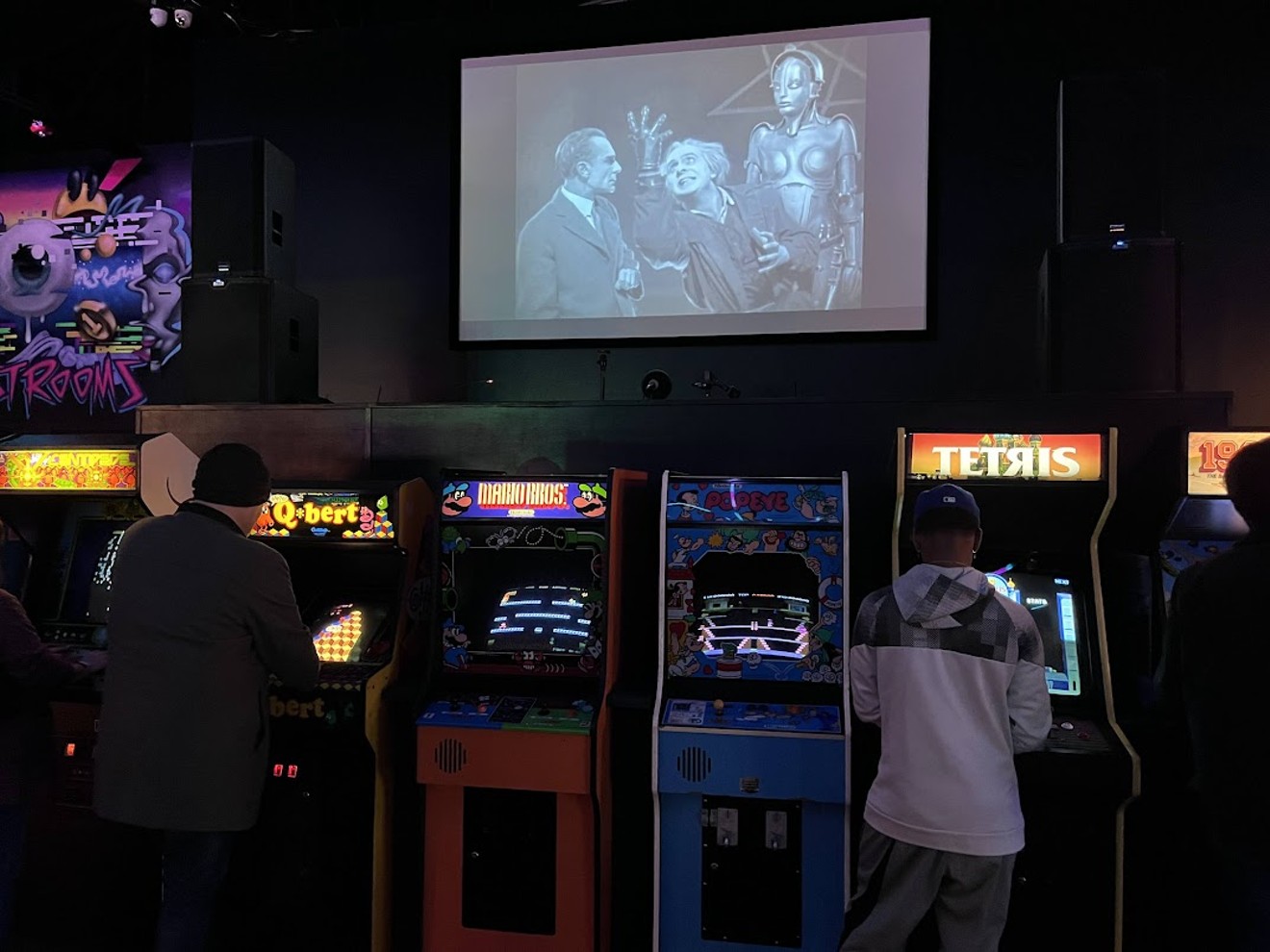 An overhead screen at Free Play Dallas in Trinity Groves plays the classic sci-fi film Metropolis as players try to get the high score on classic arcade machines.